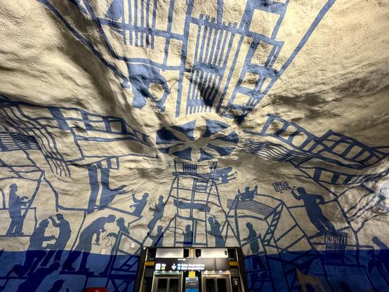 An artistic mural covers the ceiling of a subway station, featuring blue silhouettes of workers engaged in various activities. The background is textured and light-colored, creating a dynamic visual effect. Subway doors are visible in the foreground, adding context to the urban environment.