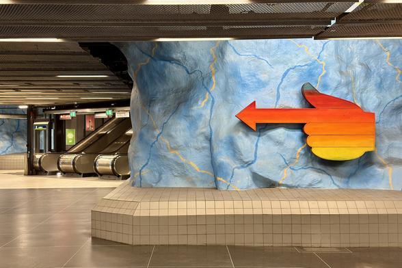 An underground station featuring a stylized blue rock wall with an artistic arrow shape in red and orange pointing to the left, alongside escalators and signage in the background.