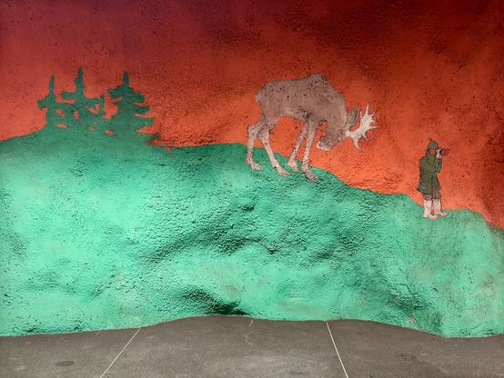 A colorful mural depicting a moose on a hill, with a person dressed in green observing it through binoculars. The background features red and green hues with silhouettes of trees.