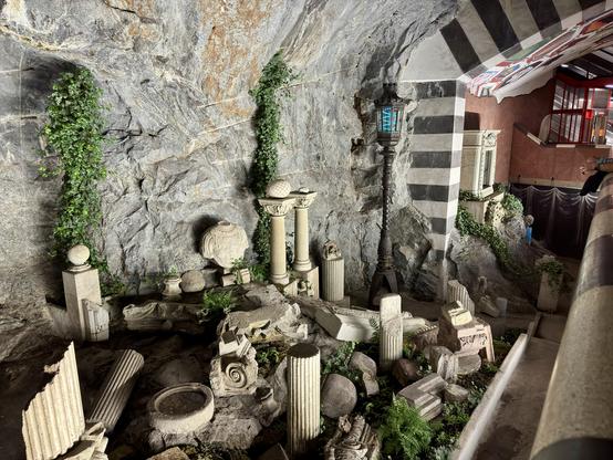 An indoor space with a rocky wall featuring various ancient-style stone sculptures, columns, and decorative elements. Green ivy and ferns sprout among the ruins, while a lantern adds a decorative touch. The ceiling has a striped design.