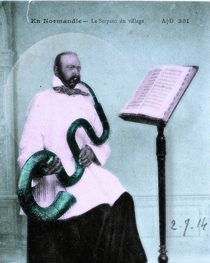 Ancienne carte postale colorisée ou l'on voit un homme barbu, en tenue d’Église, assis devant un pupitre sur lequel est posée une partition. Il tient dans ses mains et à sa bouche un long instrument sinueux formant deux boucles, il a les joues gonflées d'air et semble souffler dans l'instrument. La mention tapuscrit précise en haut de la photographie "En Normandie - le serpent du village".