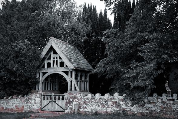 monochrome shot of roofed wooden church gateway in long stone wall,  surrounded by trees