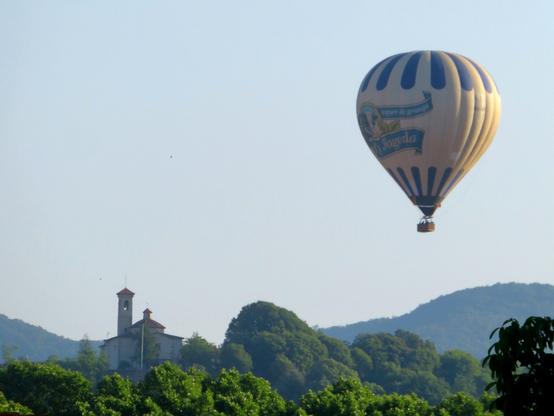 The image features a hot air balloon floating in a clear blue sky. The balloon is predominantly white with blue vertical stripes and displays the text "La Fageda" along with a logo. Below the balloon, there is a landscape with lush green trees and a building with a bell tower, likely a church, situated on a hill. The background includes rolling hills and a distant mountain range, creating a serene and picturesque scene. The overall composition highlights the contrast between the balloon and the natural surroundings.

Provided by @altbot, generated privately and locally using Ovis2-8B

🌱 Energy used: 0.126 Wh