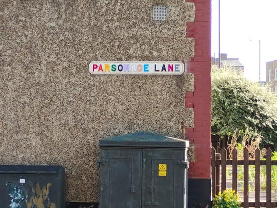Street sign for Parsonage Lane where the individual letters have been painted in the colours of the Pride rainbow -- red, orange, yellow, green, blue, violet, white, pink, light blue, brown, black, purple, grey.