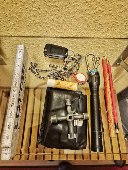 Auf einer Glasplatte liegen ein Gliedermaßstab, ein Universalschlüssel, ein Geldbeutel, ein Zimmermannsbleistift, ein Bleistift, eine Kette mit Schlüsselbund, eine kleine Taschenlampe und ein Lippenpflegestift. Alles liegt bunt durcheinander, und das Bild wurde von oben aufgenommen.