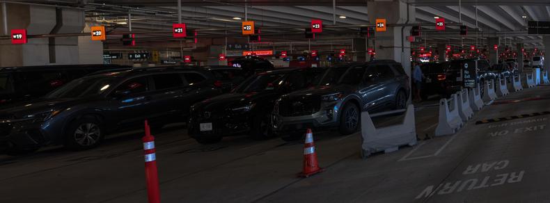 Brightly-illuminated numbered signs, whose backgrounds alternate between red and orange, hang down from the  ceiling of a large parking garage.  The numbered signs appear smaller as they continue to appear  in rows into the distance.  Parked cars loom dark, and exhibit curious geometric distortions.