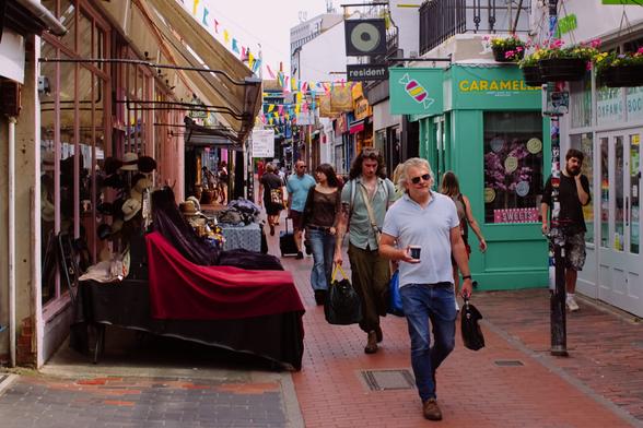 A bustling pedestrian street scene filled with life and color. People walk casually along a narrow, brick-paved lane lined with independent shops. On the left, vintage hats and garments are displayed under cream-colored awnings. On the right, pastel-colored storefronts include a sweet shop called “Caramella” and an Oxfam bookshop. Bright bunting stretches overhead, adding a festive touch. In the foreground, a grey-haired man in sunglasses walks confidently with coffee in hand. Behind him, a man with long curly hair and tattoos carries shopping bags. The street feels vibrant, eclectic, and full of everyday stories unfolding in a charming urban setting.