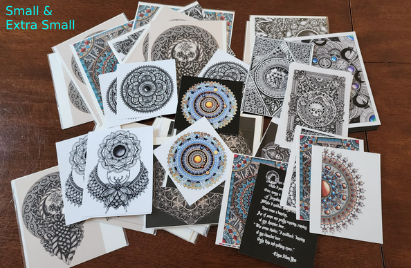 A jumble of small prints scattered on a table; a mixture of black ink line art, colour mandalas, celtic knotwork, various designs