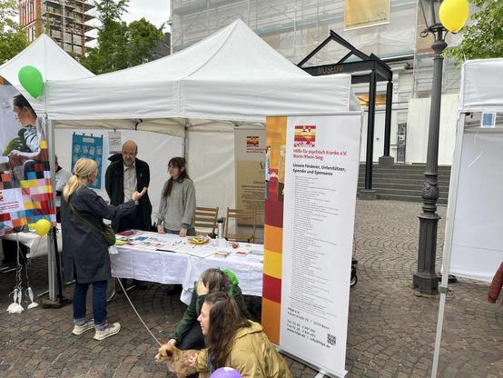 Zu sehen ist ein Foto des Aktionsstandes des HfpK e.V. Bonn/Rhein-Sieg auf dem zurückliegenden Selbsthilfetag in Siegburg. Zwei Personen aus dem Verein stehen hinter dem Infotisch und reden mit einer Besucher*in.