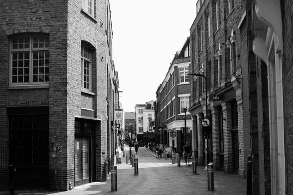 This black and white photograph captures a quiet, paved street in an urban setting, likely Shoreditch, London, looking down its length towards a brighter horizon. Tall brick buildings line both sides, with intricate window frames visible. On the left, a multi-story building of darker brick has large, gridded windows and visible signs of age. The right side features more varied architecture, including a building with columns at ground level and a prominent "tala" sign. Several bollards run down the centre of the cobbled street, leading the eye into the distance where more buildings are faintly visible under a bright, overcast sky. A few indistinct figures can be seen walking further down the street.