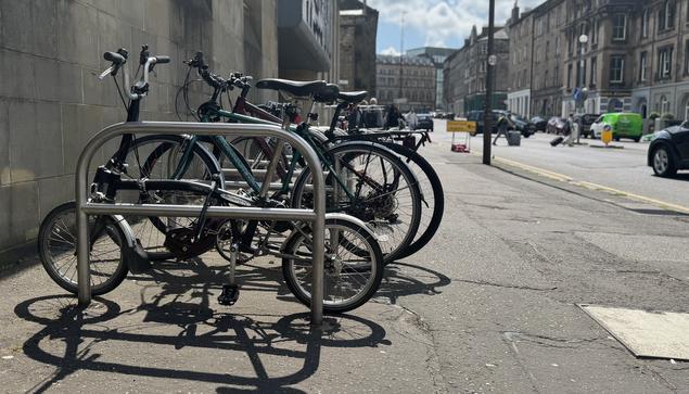 A set of Sheffield cycle racks are sited outside a sandstone building on a sunny Edinburgh street. The sun is high and the various cycles parked cast a long shadow towards the camera - a black and silver folding bike nearest the camera, with four other bikes parked up beside it. In the background, tall tenement buildings lead towards the horizon line with blue sky beyond, while the background of the image shows a handful of cars and pedestrians moving through the space.