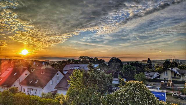 Sonnenuntergang über einer friedlichen, ländlichen Landschaft mit roten Hausdächern und grünen Wiesen. Dramatische Wolken am Himmel reflektieren warme Gelb- und Orangetöne der untergehenden Sonne. Im Hintergrund sanfte Hügel mit Bäumen und Gebäuden.