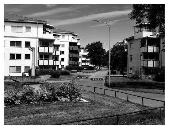 En svartvit liggande bild på en gång- och cykelväg mellan två rader av flerfamiljshus. Raden till vänster lyser vit i solskenet.
