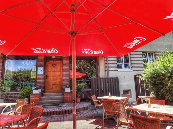 Terrasse eines Eiscafés mit Tischen und Stühlen. Über allem ein roter Sonnenschirm, der Schatten spendet. Im Zentrum des Bildes Stufen, die zum Eingang des Cafés führen.