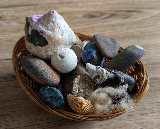 A high-angle shot shows a small, woven, light brown wicker basket filled with a variety of natural treasures on a light wooden surface. The collection includes several smooth, grey and brown pebbles, a prominent white, irregular-shaped stone with hints of purple and black, a small, round, white stone with a crack, and a vibrant blue and black, angular stone. Also visible are a white and dark mussel shell, a small, ridged, light brown scallop shell, and a small clump of light-coloured wool fibres. The wooden surface has a visible grain.