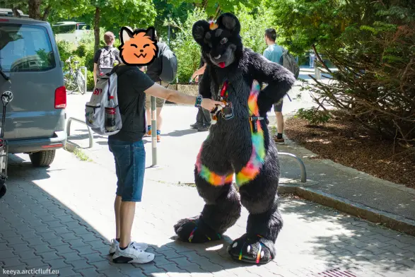 Photo of me in black jaguar fursuit, standing in a parking lot. A friend is booping the nose of a fox plush that I'm wearing on a lanyard around my neck, while I'm standing there with my hands on my hips, looking offended.