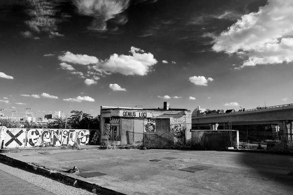 Black-and-white photo of a small kiosk, perhaps the shop of a gas station that existed earlier in this location. The building and the walls next to it are covered in graffiti. The sky is wide, with some clouds.