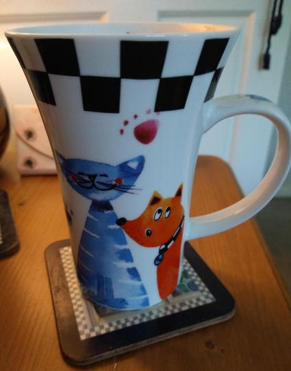 Large mug of tea on a bedside cabinet with a checkerboard pattern at the top.
A large blue cat is on the left and a small orange dog on the right