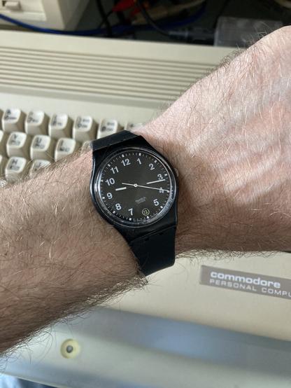 A black 1998 Swatch wrist watch with a white typeface and hands on my left wrist. A Commodore 64C is visible in the background.