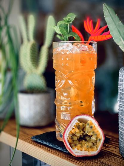 bright orange cocktail with mint garnish and swizzle stick sitting on a black coaster next to a half passion fruit