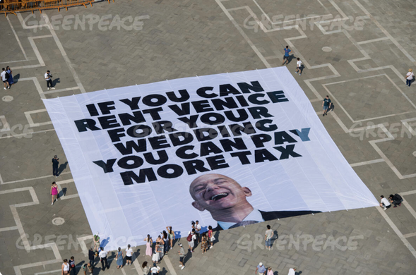 A huge banner on the floor of Piazza San Marco, Vienna, Italy.
A photo of a laughing Jeff Bezos with the text 'It you can rent Venice for your wedding you can pay more tax.'