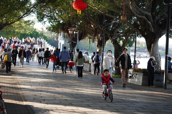 kid on a bicycle is riding over the pavement, a crowd of people can be seen in the distance. The street is surrounded by trees. The kid is wearing a red jacket and his bike is also red.