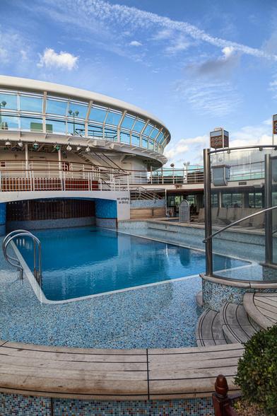 We're on the top deck of a cruise ship, standing at one corner of a swimming pool and looking diagonally across to the end of the pool which resides within an alcove underneath a bit of deck above it and a circular sun deck shielded by glass panels above that. There's nobody around at all and the ship is at anchor so the pool water is flat with a hint of vibration on its surface either from a small breeze or the ship's engines. The surrounding area of the pool is mosaic-tiled in shades of blue and the day has a cool blue sky with some puffy white clouds. It all feels quiet, chilly, peaceful. Some wooden steps leading to the pool are to the right and there's part of a glass shower in view too.