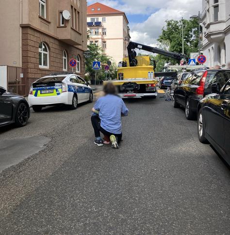 Polizei-Streifenwagen neben einem Abschleppwagen mit ausgefahrenem Arm in einer Wohnstraße, auf der Straße kniet ein Erwachsener neben einem Kind.
