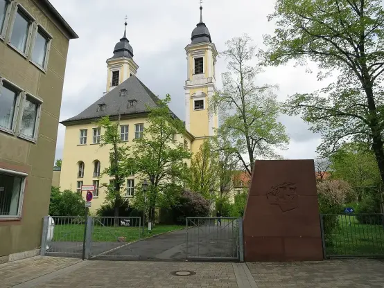 The image shows a building with two towers, likely a historical or significant structure, surrounded by trees and a gate. There’s also a large sculpture or monument in front of the building, positioned near the entrance path. The sky is cloudy, which adds to the overall ambiance of the scene. The architecture appears to be a combination of traditional styles, and the area looks well-maintained with greenery.

Image Credits: Wikimedia / Didi43 / CC BY-SA 4.0