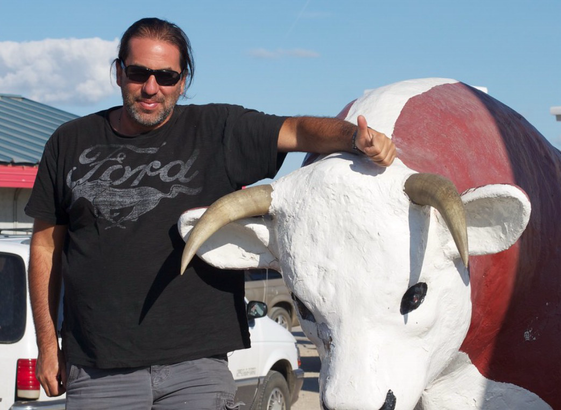 Man in dark glasses and worn t-shirt rests arm with thumbs up sign on a statue of a large bull.