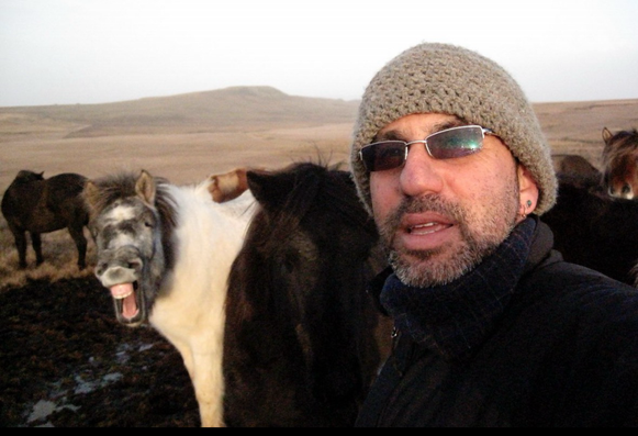 Man in dark glasses wearing a knit cap standing in front of a long haired horse making what looks like a laughing expression.