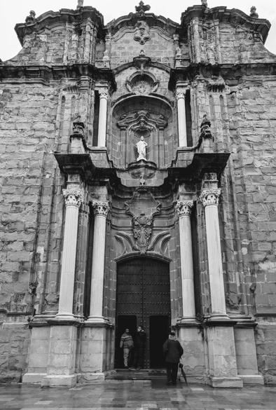 A dramatic low-angle, black and white shot captures the ornate facade of a grand stone church, the Iglesia de San Mateo in Tarifa, Spain. The imposing entrance features large, dark wooden double doors, with two figures standing on the steps, and a third figure holding an umbrella walking away from the entrance, suggesting light rain. Flanking the doorway are two pairs of majestic Corinthian columns, supporting an elaborate pediment. Above the entrance, a niche houses a statue, while the entire front is constructed from rough-hewn, large stone blocks. The intricate detailing of the baroque architecture is highlighted by the monochrome tones.