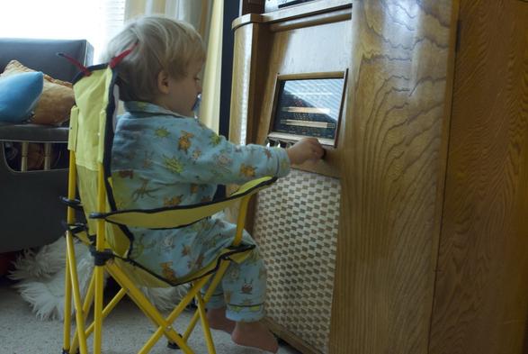 A photo of 2-year old child in pajamas sucking on a pacifier sitting in a little yellow chair turning the knobs on an old wooden valve radio that's much bigger than she is.