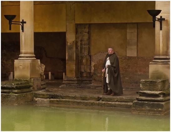 A figure dressed in a brown cloak stands near a greenish ancient pool, surrounded by stone columns and walls in a historical setting. The architecture suggests a Roman or classical influence.