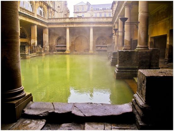 An outdoor Roman bath with green water, surrounded by stone columns and arches. Mist rises above the water, creating a serene atmosphere. The architecture features ornate details, blending historical elements with a tranquil setting.