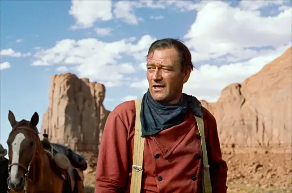John Wayne, en primer plano, viste una camisa roja y tirantes amarillos, con un pañuelo oscuro alrededor del cuello. Su expresión es seria y contemplativa. Al fondo, se aprecian imponentes formaciones rocosas desérticas bajo un cielo parcialmente nublado. Un caballo pardo con una silla de montar está ligeramente desenfocado detrás de él. La escena evoca un ambiente del oeste americano.