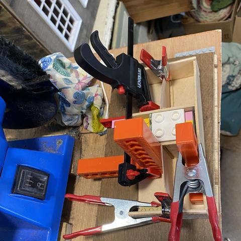 A cluttered workbench scene featuring various clamps, including a black and red clamp, multiple red spring clamps, and orange blocks and white and yellow Lego-brick pieces. There is also a blue bench polisher, in the background and the corner of the dolls house in the left top. A cloth for wiping excess glue also sits on the workbench.