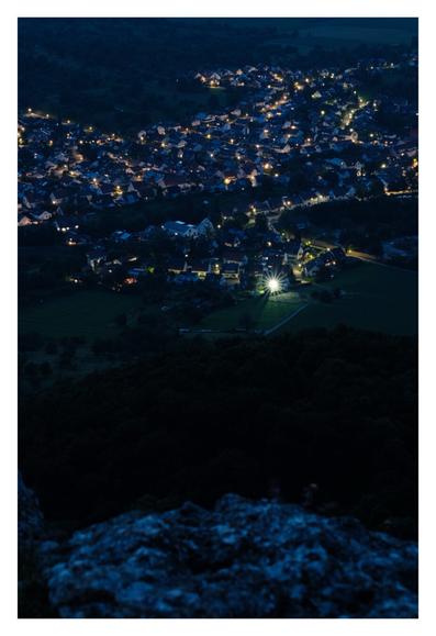 Foto im Hochformat. Blick von weit oben auf eine Stadt bei Nacht. Im Vordergrund ragt ein Felsen ins Bild. Unten im Tal sind die Lichter einer Ortschaft. Häuser, in deren Fenstern Licht brennt. Relativ weit vorne ist ein besonders helles Licht, welches wie ein Stern mit Strahlen leuchtet.