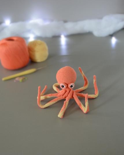 Un poulpe crocheté en corail et jaune pastel est posé avec les tentacules relevées sur un fond gris avec en fond 2 pelotes de la même couleur que le poulpe, un crochet et des anneaux marqueurs. En arrière plan, il y a une guirlande lumineuse et un nuage de ouate.