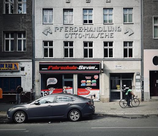 The image features a street view of a gray building with the text "PFERDEHANDLUNG OTTO-MASCHE" engraved above the windows. Below, a driving school named "Fahrschule-Sevim" is prominently displayed.