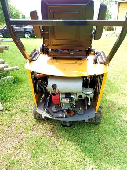 Rear of a a mini-excavator with the back removed. A red and silver engine sits in there, a large exhaust muffler with glass wrapping at the top, a hydraulic pump on the right, a small combustion and large cooling air intake on the left. Some cables and rubber mounts on the bottom.