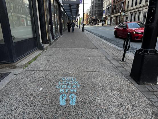 Blue stencilled text saying you look great btw is written on a sidewalk on Barrington Street, Halifax, NS. A red car is stopped on the street and two people walk toward the camera
