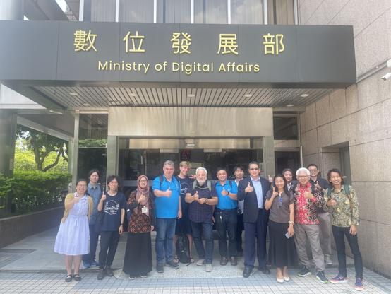 Eine Gruppe von 15 Personen steht vor dem Eingang des „Ministry of Digital Affairs“ in Taiwan. Über dem Eingang sind große Schriftzeichen in traditionellem Chinesisch sowie die englische Bezeichnung der Behörde angebracht. Die Personen – darunter Mitglieder der LibreOffice-Community und Delegierte der The Document Foundation – lächeln in die Kamera und zeigen Daumen-hoch-Gesten. Einige tragen LibreOffice-T-Shirts. Es ist ein sonniger Tag, im Hintergrund sind Glasfassaden und Grünpflanzen zu sehen.
