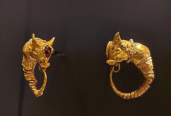 Two gold earrings, the finials are shaped like bull heads, with intricate detailing and coiled lower sections. Red gemstone set in the forehead of the animals. The earrings are displayed against a dark background.