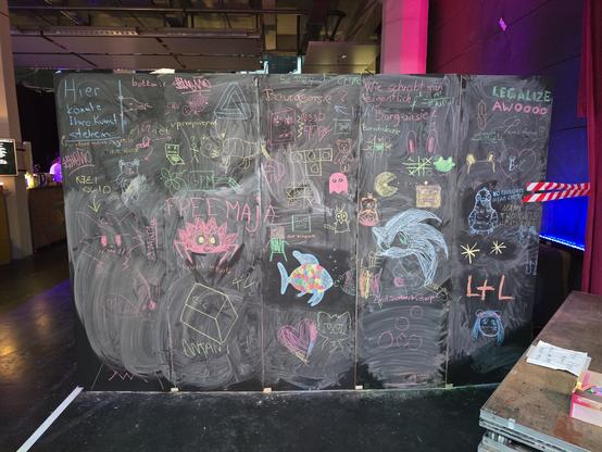 Eine ca. 4 x 2 m große Kreidetafel mit bereitliegenden Kreidestiften. Oben links steht in einem Kasten: "Hier könnte Ihre Kunst stehen"
Darunter hat jemand in andere Farbe geschrieben "Hier stand meine Kunst ;(". Noch wer anders hat in Anführungszeichen "Ihre Kunst" daruntergesetzt.

Es gibt allerlei Geschriebenes, z.B. "Wie schreibt man eigentlich Burgwasie?" Jemand hat "Burschhase" geantwortet, wer anders "Bourgeoisie?"

Da steht "Legalize Awoooo",  "Free Maja", "Rewrite in Rust!", "bottom :3" und mehr.
Ins Auge fallen vor allem die gezeichneten Figuren: Da wäre das Krebs-Maskottchen von Rust mit großen Kulleraugen, ein Regenbogenfisch, der einen anderen bunten Fisch zu küssen scheint, der Weltraumigel Sonic (mutmaßlich), Teddybären, Katzen, Katzengesichter, Robotergesichter, eine Taube, ein Bärchen mit Frucht auf dem Kopf, Pacman, Pacman-Geist, Figur aus Among Us, ein Hund, ein geometrisch unmögliches Dreieck, irgendwas zwischen Vogel und Biber und noch einige Objekte mehr.