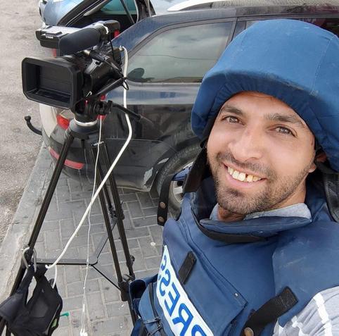 The image shows a person wearing a blue protective vest with the word "PRESS" written on it, indicating they are a journalist or media worker. The individual is smiling and appears to be taking a selfie with a professional video camera mounted on a tripod. The camera is black with a large lens and a microphone attached. The person is wearing a blue hooded jacket, which is part of their protective gear. In the background, there is a dark-colored car parked on a paved surface. The scene suggests that the person is in a professional setting, possibly preparing to film or report on an event.