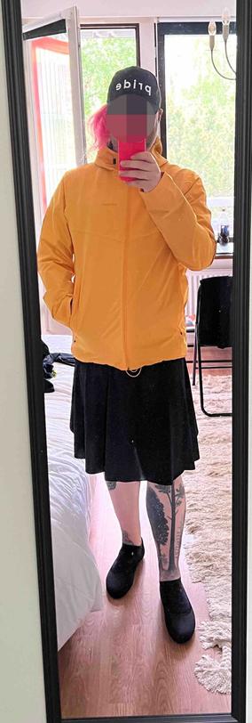 A person taking a mirror selfie. They are wearing a black base cap with the white letters “pride”, a dark yellow coat and a knee long black skirt with ballerinas. The legs are bare and tree tattoos are visible.