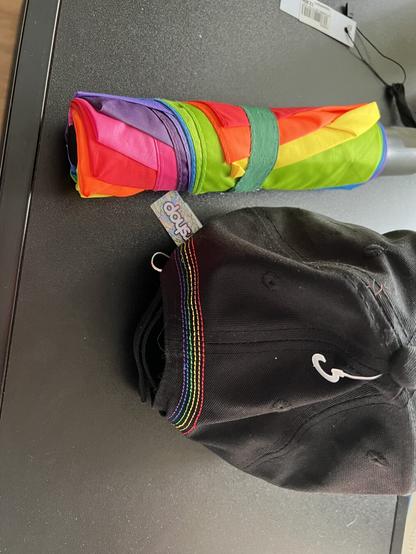 A folded umbrella with rainbow colored fabric and the backside of a black base cap with rainbow stitching