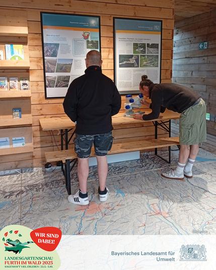 Ein Mann steht vor einer Holzwand und betrachtet ein Plakat, ein anderer steht daneben und sieht durch ein Mikroskop.