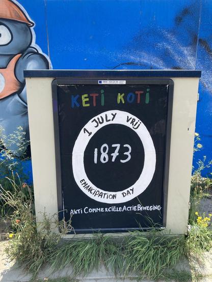 blauwe muur met graffiti, met elektriciteitskastje met reclame bord bewerkt in:
in blauw, geel, rood, groene letter KETI KOTI
In witte cirkel op zwarte achtergrond met zwarte letter boven: 1 juli vrij en onder: emancipation Day. In het zwarte midden met witte letters 1873 en onderaan het bord Anti Commerciele Actie Beweging. Wat gras omringd het bord